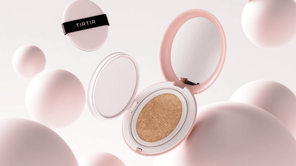 Bilden visar en öppen cushion foundation med märket "TIRTIR" på locket. Produkten är placerad mot en bakgrund av abstrakta vita och rosa sfärer som skapar en mjuk och ren atmosfär, vilket antyder en skönhetsprodukt med en elegant och modern design.
