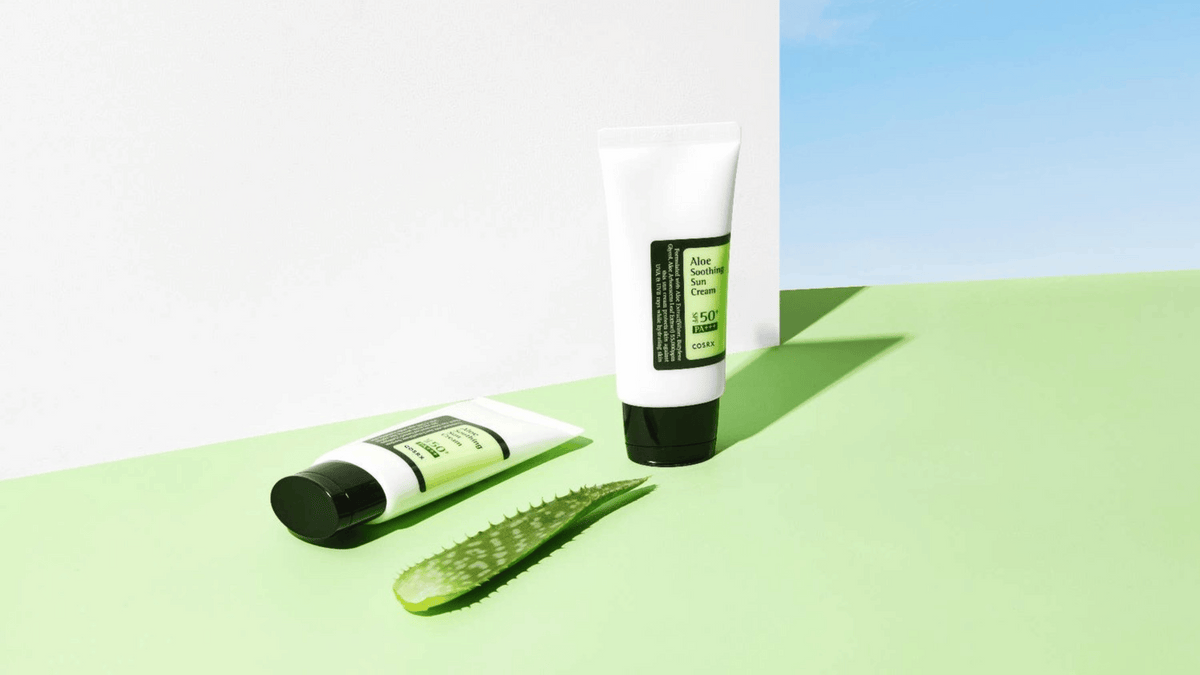 Två tuber med COSRX Aloe Soothing Sun Cream SPF50+ PA+++ står mot en ljusgrön bakgrund tillsammans med en bit färsk aloe vera och blå himmel i bakgrunden.