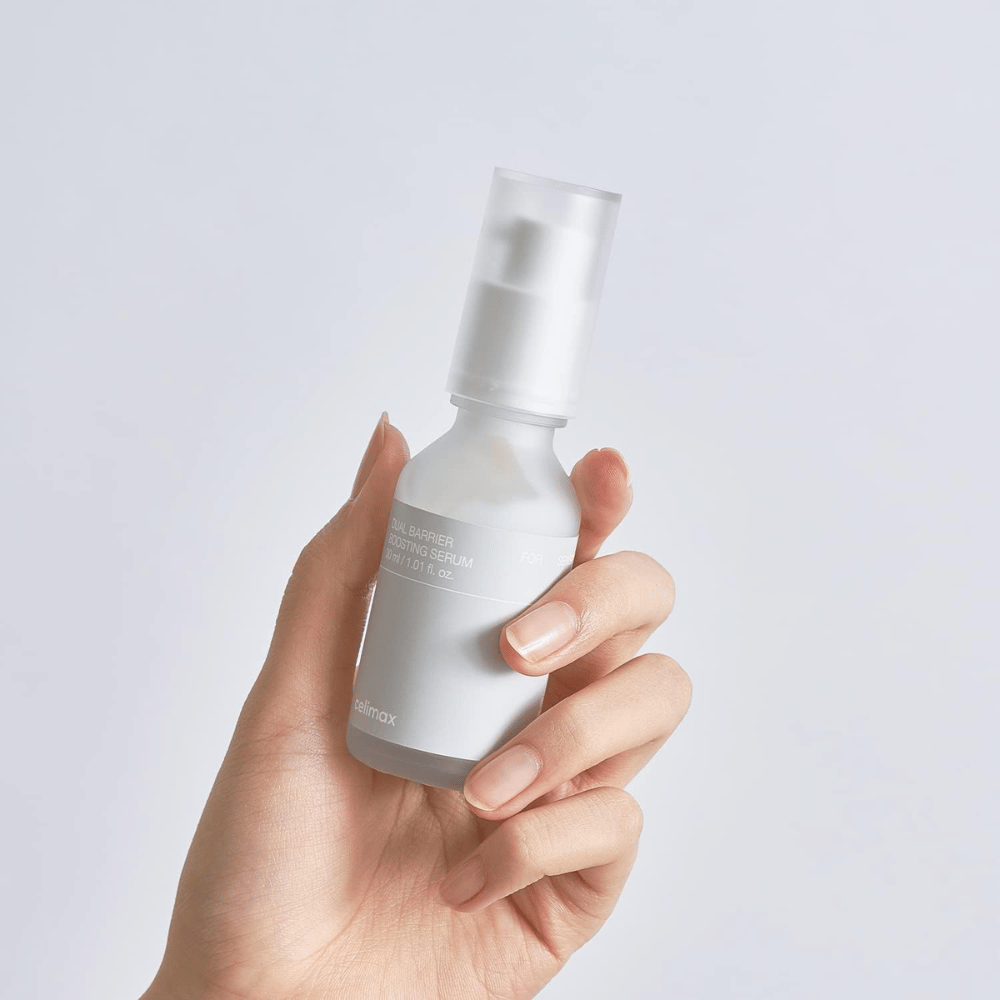 Celimax Dual Barrier Boosting Serum kädessä, minimalistinen pakkaus, joka esittelee suojamuuria vahvistavaa korealaista seerumi herkälle iholle.