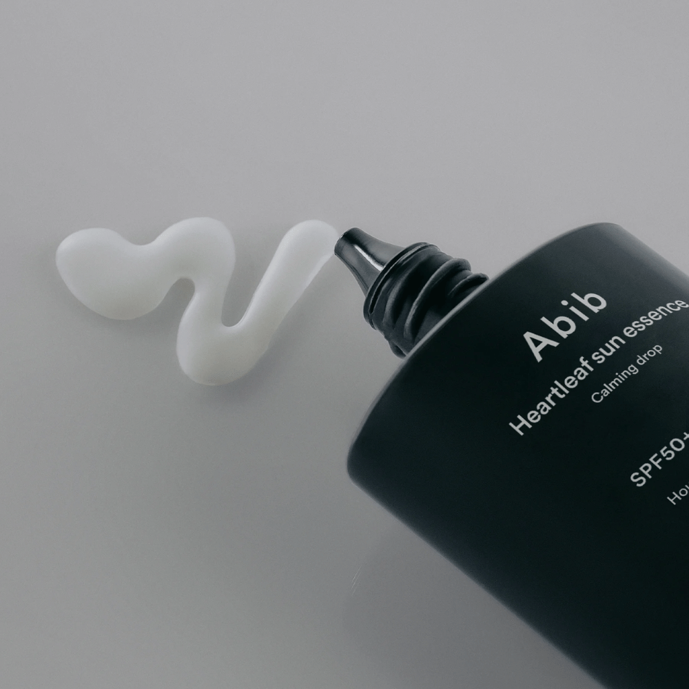 Lähikuva Abib Heartleaf Sun Essence Calming Drop aurinkovoiteesta puristettuna valkoisena, aaltoilevana viivana vaalealla pinnalla.